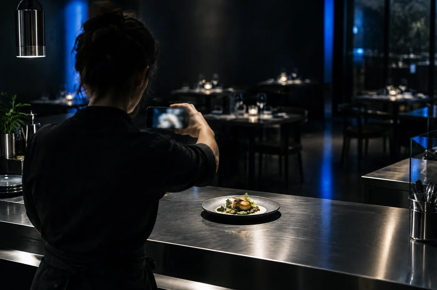 Une restauratrice vue de dos photographie une assiette dressée sur le comptoir de sa salle